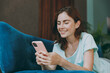 © ViDi Studio - Young happy smiling cheerful fun woman she 20s in casual clothes mint t-shirt hold in hand use mobile cell phone lying on blue sofa indoor rest at home in own room apartment People lifestyle concept.