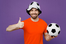 Young Excited Happy Fan Man He 20s Wears Orange T-shirt Cheer Up Support Football Sport Team Hold In Hand Soccer Ball Panama Hat Watch Tv Live Stream Show Thumb Up Isolated On Plain Purple Background.