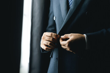 Confident businessman buttoning or adjust classic blue suit near window in hotel room at the morning. Handsome man wearing a nice suit on wedding day.