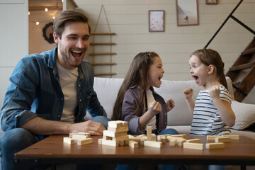 Wall Mural - Joyful father with daughters plays at home. Kids remove wooden blocks from tower. Board game.