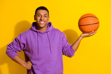 Sticker - Portrait of attractive cheerful sportive guy fan holding on palm orange ball isolated over bright yellow color background