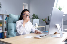 Sick Asian Female Office Worker, Has Cold And Flu, Coughs And Sneezes Into Tissue, Works At Computer.