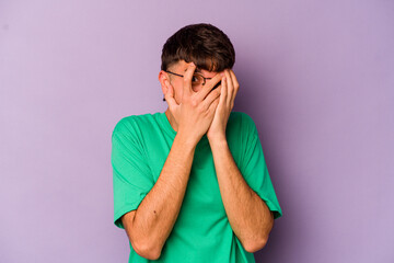 Wall Mural - Young caucasian man isolated on purple background blink through fingers frightened and nervous.