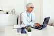 © Jelena Stanojkovic - A mature women doctor sitting at the workplace and looking at the laptop during the day.  Portrait of a female doctor wearing white uniform using laptop to talk to patient online.