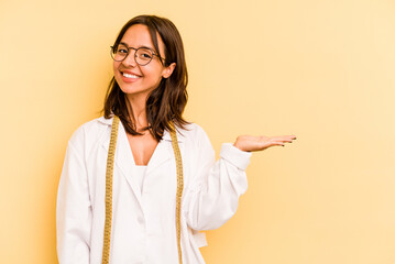 Young nutritionist hispanic woman isolated on yellow background showing a copy space on a palm and holding another hand on waist.