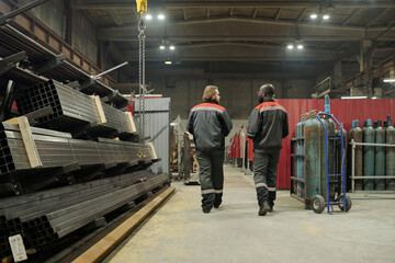 Poster - Rear view of two young interracial male workers in uniform discussing quality of new equipment while moving along aisle in warehouse