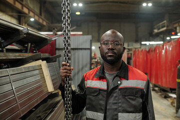 Aufkleber - Young serious black man in workwear holding by bunch of thick metallic chains while checking adjustments of new industrial equipment