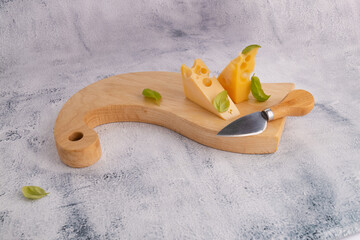 Wall Mural - Maasdam cheese on a wooden board with basil leaves and a knife. Cheese cubes on a board on a gray stone table with copy space 