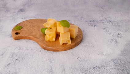 Wall Mural - Maasdam cheese on a wooden board with basil leaves. Cheese cubes on a board on a gray stone table with copy space	