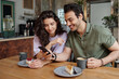 © pressmaster - Young couple looking through funny photos or video in smartphone held by guy while having cappuccino or latte with cheesecakes
