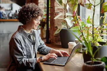 Young serious businesswoman or freelancer in casualwear sitting by window in front of laptop in cafe and typing while working