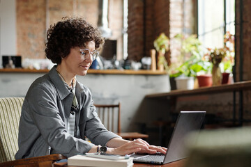 Poster - Young female freelancer or owner of cafe in casualwear and eyeglasses sitting in front of laptop by table and organizing work