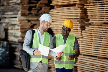 Black worker and warehouse inspector cooperate while analyzing paperwork at lumber department.