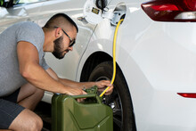 A Young Man Steals Gasoline From The Gas Tank In The White Car. A Young Man Pumps Gasoline From A Gas Tank Into A Canister. Fuel And Oil Crisis. The Concept Of Gasoline Prices And The Oil Crisis.