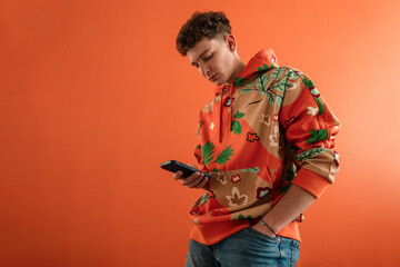 Wall Mural - Fashion studio portrait of a young man in hoodie with smartphone posing over orange background.