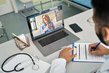 Over Shoulder View Of Videocall Chat With Female Older Patient And Indian Male Doctor In Modern Clinic Hospital Using Laptop Computer, Consulting Online Remotely. Telemedicine Healthcare Concept.
