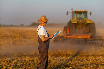 An elderly farmer with a digital tablet in his hands walks through the field, overseeing the plowing of the wheat field after harvest . Tractor in the background.