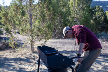 Man in sweatshirt investigating a outdoor propane camping stove to cook food at camp site in Utah in early morning.