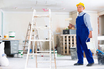 Wall Mural - Man painter in coverall and hardhat carrying bucket and paint roller. Male builder working in construction site.