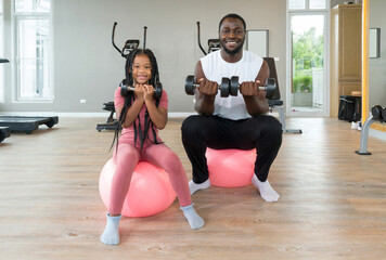 Young short curly black hair man and his daughter lifting barbell with both hands while sitting on yoga ball. Happy family enjoy holiday together in fitness center.