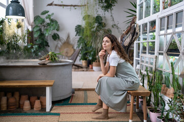 Wall Mural - Calm woman sits on bench in home garden surrounded plants thinking, looking out the window. Thoughtful gardener female resting after work enjoying her greenhouse. Doubtful italian lady in floral shop