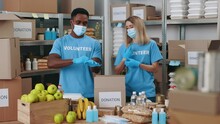 Two Young People Putting On Medical Face Masks And Rubber Gloves While Standing Together At Food Bank. Multiracial Volunteers Working At Warehouse For Helping People During Coronavirus.