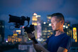 Man filming with camera and gimbal. Portrait of videographer against illuminated urban skyline at night..
