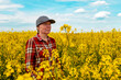© Bits and Splits - Confident and self-assured farm worker wearing red plaid shirt and trucker's hat standing in cultivated rapeseed field in bloom and looking over crops