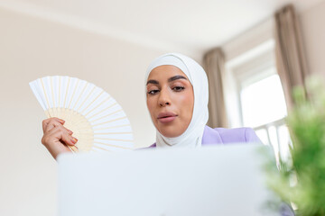 Tired overheated young Arabic  woman hold wave fan suffer from heat sweating indoor work on laptop at home office