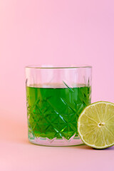 Wall Mural - green alcohol drink in glass near half of fresh lime on pink.