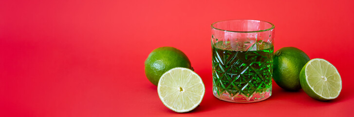 Wall Mural - green alcohol drink in faceted glass near sour limes on red, banner.