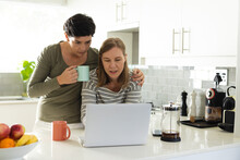 Caucasian lesbian woman holding coffee and looking at laptop being used by girlfriend in kitchen