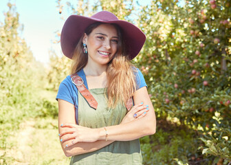 Wall Mural - A beautiful female farm worker standing with her arms crossed on a fruit farm during harvest season. Young happy farmer between fruit trees in summer. Agricultural industry growing produce