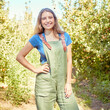 © Jade M/peopleimages.com - Portrait of a female farm worker standing on a fruit farm during harvest season. Young smiling farmer between fruit trees on a sunny day. The agricultural industry growing fresh produce