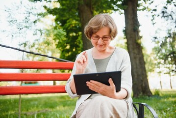 Wall Mural - Positive confident old lady posing with tablet in park. Senior grey haired woman in casual sitting on park bench and using tablet. Wireless connection concept