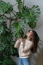 Being In Touch With Nature. Young Attractive German Woman Plant Lover Caring For Plants, Standing Near Big Monstera Houseplant And Touching Leaf, Enjoying Leisure Time By Doing Home Gardening