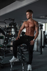  Handsome young athletic guy with hair and a bare muscular torso stands and poses near the dumbbells in the gym