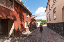 An Old Recreated Cobblestone Street With Red Craftsman Wood Houses And A Folk Costume Dressed Women, A Sunny Summer Day In Stockholm
