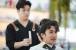 © chokniti - Asian man sitting in customer chair while getting a haircut in salon with a male hair stylist