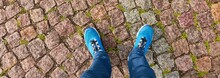 Man Standing On Cobblestone Road. Blue Jeans And Trekking Boots. Texture, Background, Wallpaper. Travel, Tourism, Walking, Cycling. Details Of Urban And Landscaping Design. Panoramic View