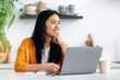 © Kateryna - Happy lovely chinese brunette young woman, creative designer, freelancer or student, sits at the kitchen with a laptop, working or study remotely, looking aside, smiling friendly, dreaming vacation