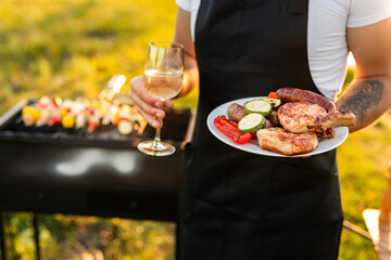 Wall Mural - Crop anonymous waiter standing in park with plate of grilled food and wineglass