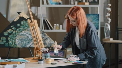 Wall Mural - Inspired busy focused redhead girl artist woman painter in art studio background paints with wide brush on canvas with acrylic oil colorful paint puts picture on easel satisfied looking at camera