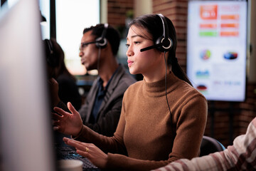 Wall Mural - Asian female receptionist working at customer service helpdesk, talking to clients on helpline support at call center. Sales consultant using remote telecommunication assistance in office.