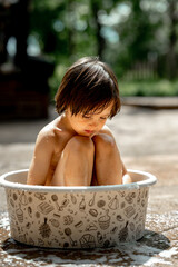 A happy Asian-looking kid with long black hair splashes in a basin in the yard of a house on a hot summer day. The concept of a happy childhood