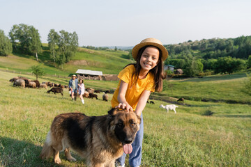 Sticker - joyful girl looking at camera near cattle dog and parents herding flock on blurred background.