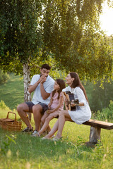 Wall Mural - happy girl sitting on bench under birch near parents playing harmonica and guitar.