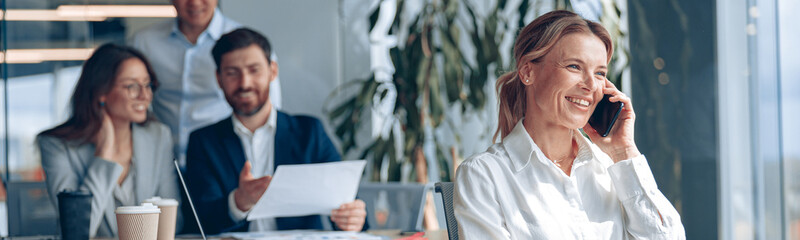Wall Mural - smiling mature lady is sitting on a chair and talking on phone during meeting in modern office.