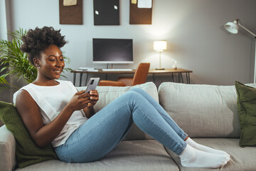 Wall Mural - Woman using mobile phone while sitting on sofa. Female is text messaging in living room. She is relaxing at home. Smiling african woman daydreaming and holding mobile phone with copy space