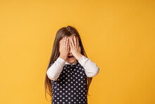Shy Child Who Is Ashamed Of His Bad Behavior Without Showing His Tears. A Little Girl Covering Her Face With Her Hands Is Afraid Of A Horror Movie On A Yellow Background. Body Language